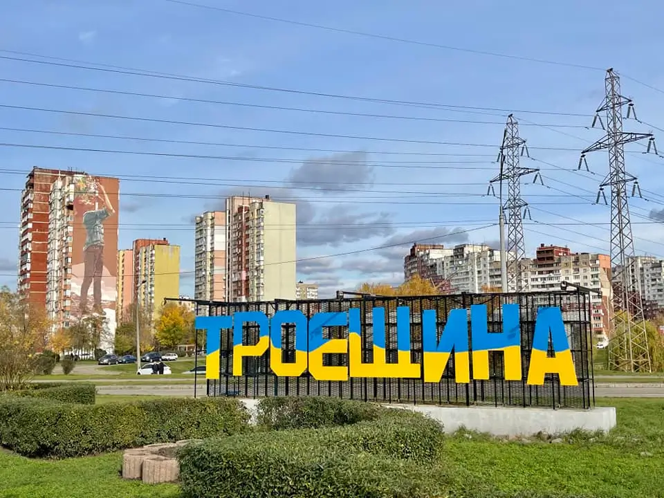 Панорамний вид Деснянського району Києва зі стелою «Троєщина» у жовто-блакитних кольорах та сучасними муралами на житлових будинках.